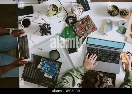 Multiple individuals engaging in electronic work on cluttered table filled with various components and tools. Hands seen handling devices and eating, Stock Photo