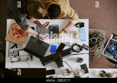 Overhead view of two people collaborating on repairing computer hardware with various tools and devices spread out on table in office setting Stock Photo