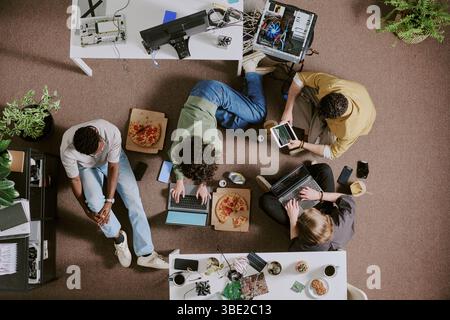 Group of diverse individuals sitting around table, working on various tech devices and eating pizza in modern office setting with greenery and tech eq Stock Photo