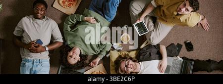 Group of young professionals lying on floor, collaborating on various projects with devices and snacks around. Multiracial team engaging in creative t Stock Photo