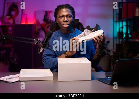 Portrait of African American man holding sneaker while sitting at desk, demonstrating product for viewers on video. He is in studio with microphone setup and laptop Stock Photo