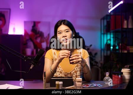 Portrait of young Asian woman showcasing beauty product on social media, in studio with various cosmetics and modern equipment, suggesting content creation and beauty tutorial Stock Photo