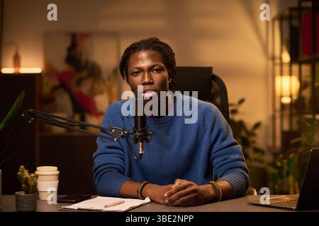 Portrait of an African American man sitting at desk, speaking into microphone, using notes, engaging in conversation. Background includes blurred abstract artwork Stock Photo