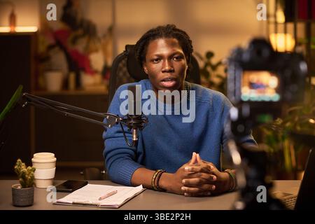 Portrait of young African American man speaking into microphone and looking at camera, while recording content in well-lit room set up for a podcast, with plants on the desk Stock Photo
