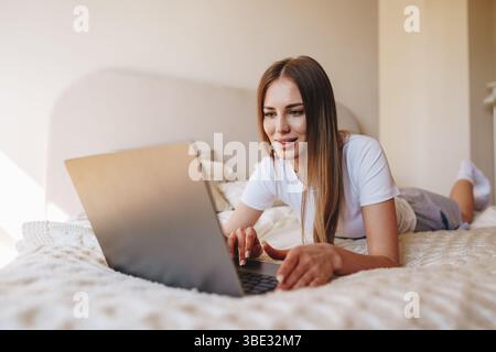 Young woman using laptop while lying on bed in cozy bedroom during the day Stock Photo