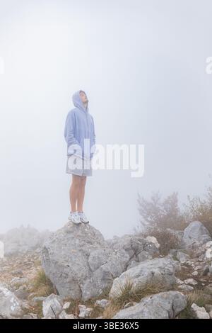 Man with hood on standing quietly top rock thick white fog surrounding background. Stock Photo