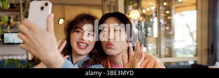 Two friends enjoy a lighthearted moment while taking a selfie in a vibrant cafe, banner Stock Photo