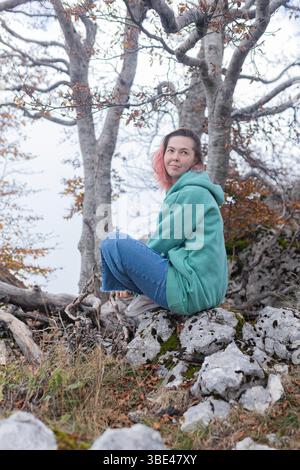 Woman with pink hair and green hoodie sitting peacefully on rocky ground under autumn trees. Stock Photo
