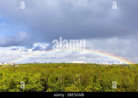 gray rainy clouds and rainbow in blue sky over green city park after rain on sunny spring day Stock Photo