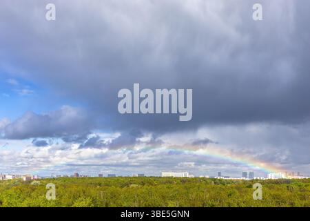 grey rainy clouds and rainbow over green city park after rain on sunny spring day Stock Photo