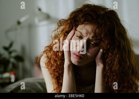 Curly-haired young woman resting her cheek on her hand, appearing deep in thought with melancholic expression in cozy indoor setting, blurred backgrou Stock Photo