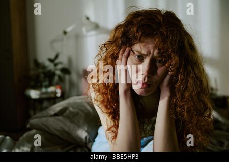 Woman with curly hair sitting indoors, resting her head in her hands, appearing distressed and deep in thought, surrounded by blurred background eleme Stock Photo