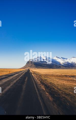 A straight dirt road stretches toward a dramatic ridge of snow-covered mountains in the Stokksnes area of Iceland, surrounded by wide open fields and Stock Photo