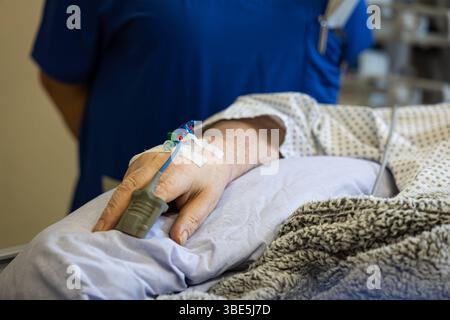 Deutschland 25. April 2025: Im Bild: Eine detaillierte Nahaufnahme zeigt die Hand eines Patienten in einem Krankenhausbett. Am Handrücken ist ein intravenöser Zugang Infusion sichtbar, gesichert mit medizinischem Klebeband. Ein blauer Schlauch führt von der Kanüle weg. Am Zeigefinger des Patienten ist ein graues Pulsoximeter angebracht, das die Sauerstoffsättigung im Blut misst. Der Patient trägt ein typisches Krankenhauskittelmuster, das im oberen Bereich des Bildes leicht sichtbar ist. Im Hintergrund ist unscharf eine Person in blauer Arbeitskleidung zu erkennen, möglicherweise medizinisches Stock Photo