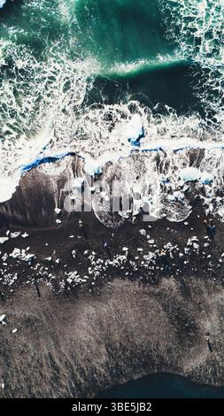 Overhead drone view of powerful ocean waves breaking over glacial ice fragments scattered across the black sand of Diamond Beach in Iceland. A stunnin Stock Photo