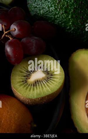 A detailed close up of fresh fruits including a sliced kiwi, dark red grapes, an avocado, and an orange on a dark background. The colorful assortment Stock Photo