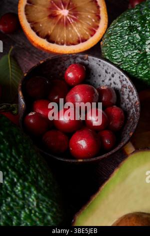 A close up of a rustic bowl filled with fresh, red cranberries surrounded by ripe avocados and citrus fruit slices on a dark wooden background, showca Stock Photo