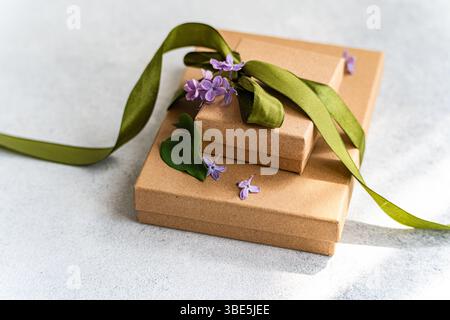 Two brown gift boxes are adorned with a green ribbon and delicate purple flowers. The elegant setup rests on a subtle backdrop, conveying warmth and g Stock Photo
