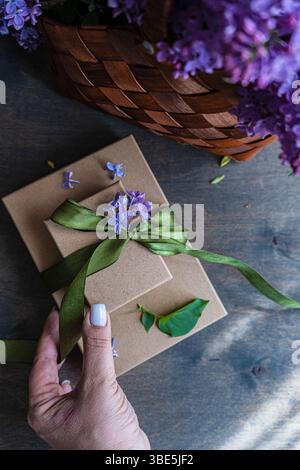A hand holds a stack of brown gift boxes tied with a green ribbon and adorned with purple flowers. A woven basket with lilacs is in the background on Stock Photo