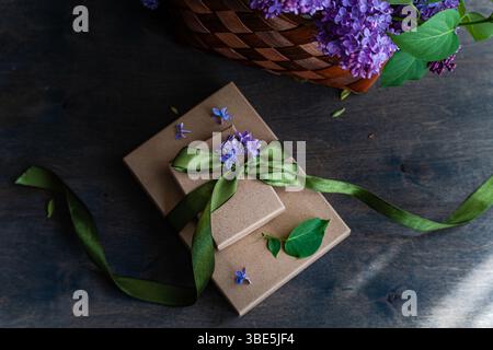 Two gift boxes wrapped in earthy paper with green ribbon and lilac flowers. They rest on a wooden surface next to a woven basket, creating a rustic, e Stock Photo