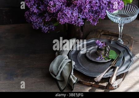 Elegant table setting with vintage plates, silverware, and delicate lilac flowers on a rustic wooden board. A soft, textured cloth napkin completes th Stock Photo