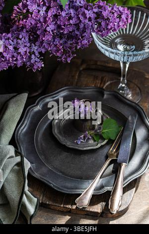 A rustic table setting features antique pewter plates and cutlery, adorned with vibrant lilac flowers in a vintage cup. A pleated napkin and glass add Stock Photo