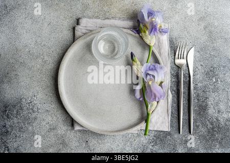 Top view of an elegant table setting featuring a concrete textured plate, clear glass, silverware, and a beautiful iris flower, arranged on a gray sur Stock Photo