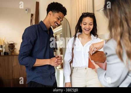 A young couple smiles while a salesperson showcases a stylish leather handbag in a modern bag store. The elegant setting and attentive customer servic Stock Photo