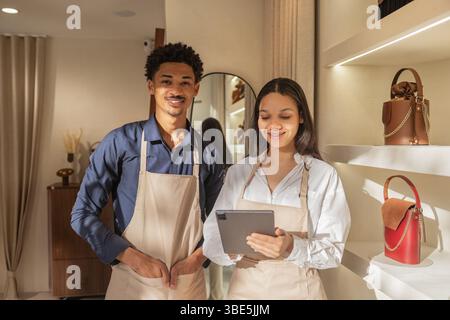 Two smiling store employees in aprons work together in a stylish, well lit bag store. They use a tablet to manage inventory and improve customer servi Stock Photo