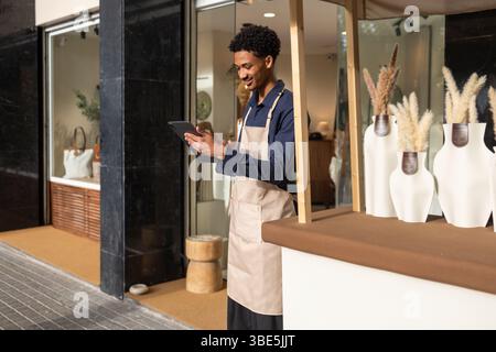 A young, smiling employee in an apron stands at the entrance of a stylish bag store, interacting with a tablet. The shop showcases elegant bags throug Stock Photo