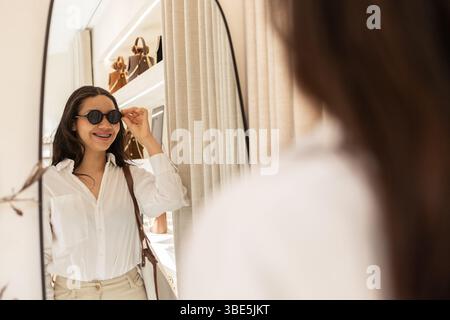 A stylish woman tries on sunglasses and smiles in front of a mirror inside a chic bag store. The shelves behind display a variety of elegant handbags, Stock Photo