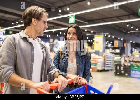 A couple enjoys shopping in a supermarket, pushing a cart full of products. They smile and engage in friendly conversation, creating a warm and welcom Stock Photo