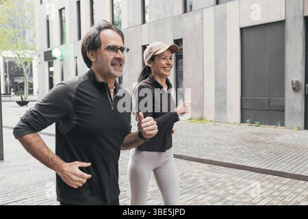 A cheerful couple enjoys a refreshing jog together on a city sidewalk, dressed in athletic wear and sharing smiles, embodying a healthy and active lif Stock Photo