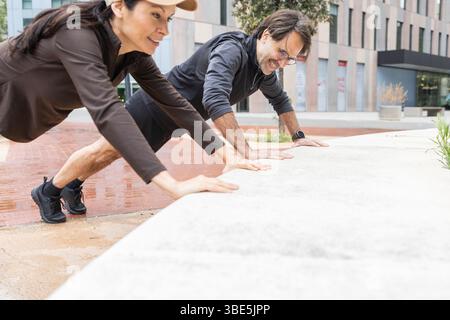 A couple is engaging in an energetic outdoor workout, doing inclined push-ups on a concrete ledge in an urban setting. They are dressed in athletic ge Stock Photo