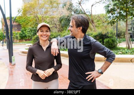 A cheerful couple takes a break during their outdoor exercise routine, standing on a scenic path surrounded by greenery. Both dressed in athletic wear Stock Photo