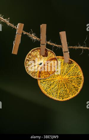 Two translucent dried orange slices are artistically suspended from a twine using wooden clothespins against a dark green background. Stock Photo