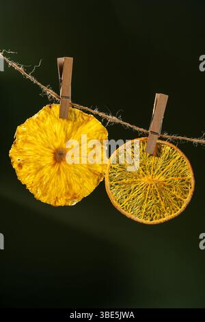Two colorful dried fruit slices, a vibrant pineapple and a translucent orange, hang from a rustic twine secured with wooden clothespins, against a dar Stock Photo