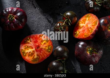 A selection of heirloom tomatoes, whole and halved, with glistening dewdrops. The dark surface and dramatic lighting create a rich contrast, highlight Stock Photo
