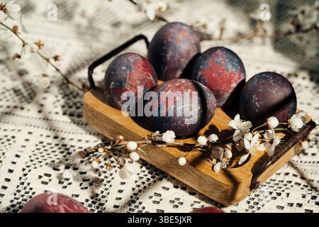 Artistic Easter eggs with a cosmic paint theme are displayed on a wooden tray surrounded by cherry blossoms, placed on a patterned crochet tablecloth, Stock Photo