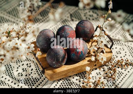 A rustic composition of four uniquely painted Easter eggs, nestled among delicate cherry blossoms on a wooden tray, lying over a textured crochet tabl Stock Photo