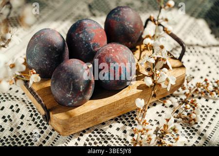 Artistic galaxy-themed Easter eggs displayed on a wooden tray, surrounded by spring blossoms on a patterned crochet tablecloth backdrop. Stock Photo