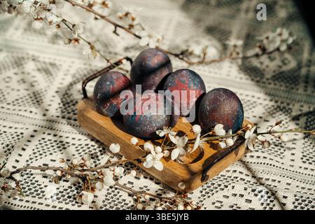 Cosmic-painted Easter eggs displayed on a rustic wooden tray, adorned with delicate white spring blossoms, set against a patterned crochet tablecloth Stock Photo