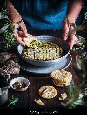 A vibrant bowl of green hummus garnished with herbs is placed on a rustic wooden table. Beside it, a variety of crackers are arranged as someone dips Stock Photo
