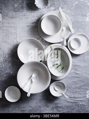 Top view of an elegant arrangement of white ceramic dishes and utensils on a textured grey background. Includes plates, bowls, and a small green sprig Stock Photo
