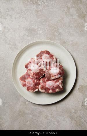 Five raw oxtail pieces are neatly arranged on a round, white ceramic plate. The marbled texture and round vertebrae are visible, showcasing fresh beef Stock Photo