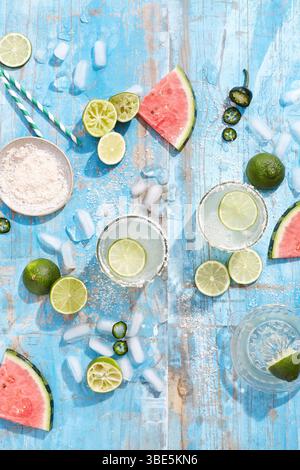 Top view of a vibrant summer cocktail arrangement featuring limes, watermelon slices, ice cubes, and sea salt scattered on a rustic blue wooden surfac Stock Photo