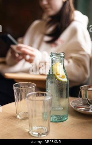 Cropped unrecognizable blurred young woman in a cafe, engaged with her smartphone near a cold water bottle with a lemon slice and empty glasses on a w Stock Photo