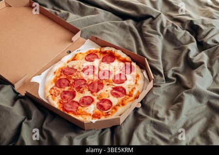 A freshly baked pepperoni pizza sits inside an open cardboard box placed on a crinkled gray bed sheets, showcasing a casual, cozy eating setting. Stock Photo