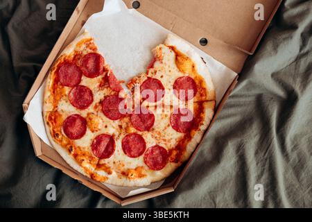 Top view of a freshly baked pepperoni pizza lying in an open cardboard box on a bed with grey sheets. Stock Photo