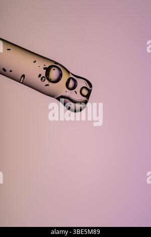 A dropper dispenses transparent serum against a soft pink background, showing bubbles and reflections within the liquid, suggesting purity and skincar Stock Photo